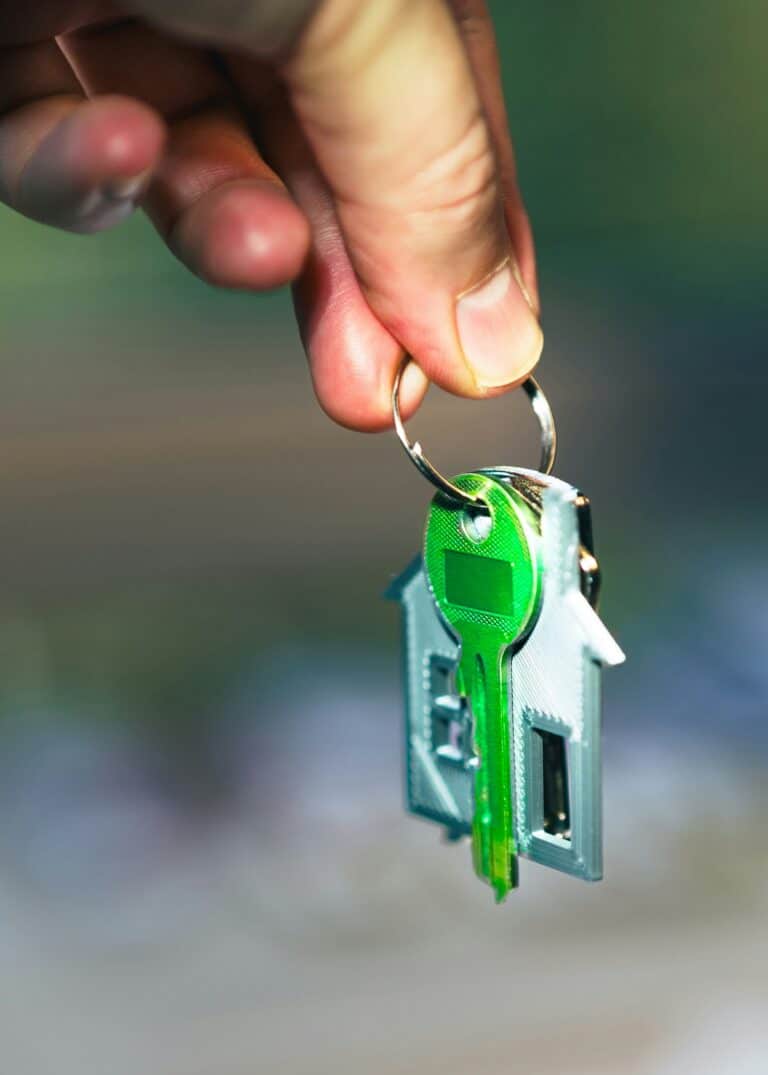 A close-up of a person holding a house key with a green accent and a house-shaped silver keychain, representing a successful senior relocation and downsizing transition.