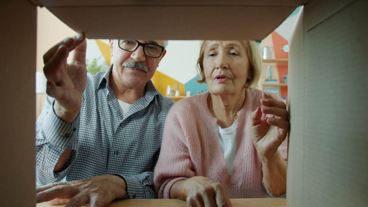 An elderly man with glasses and an older woman in a pink cardigan looking curiously inside a cardboard moving box while packing or sorting items.