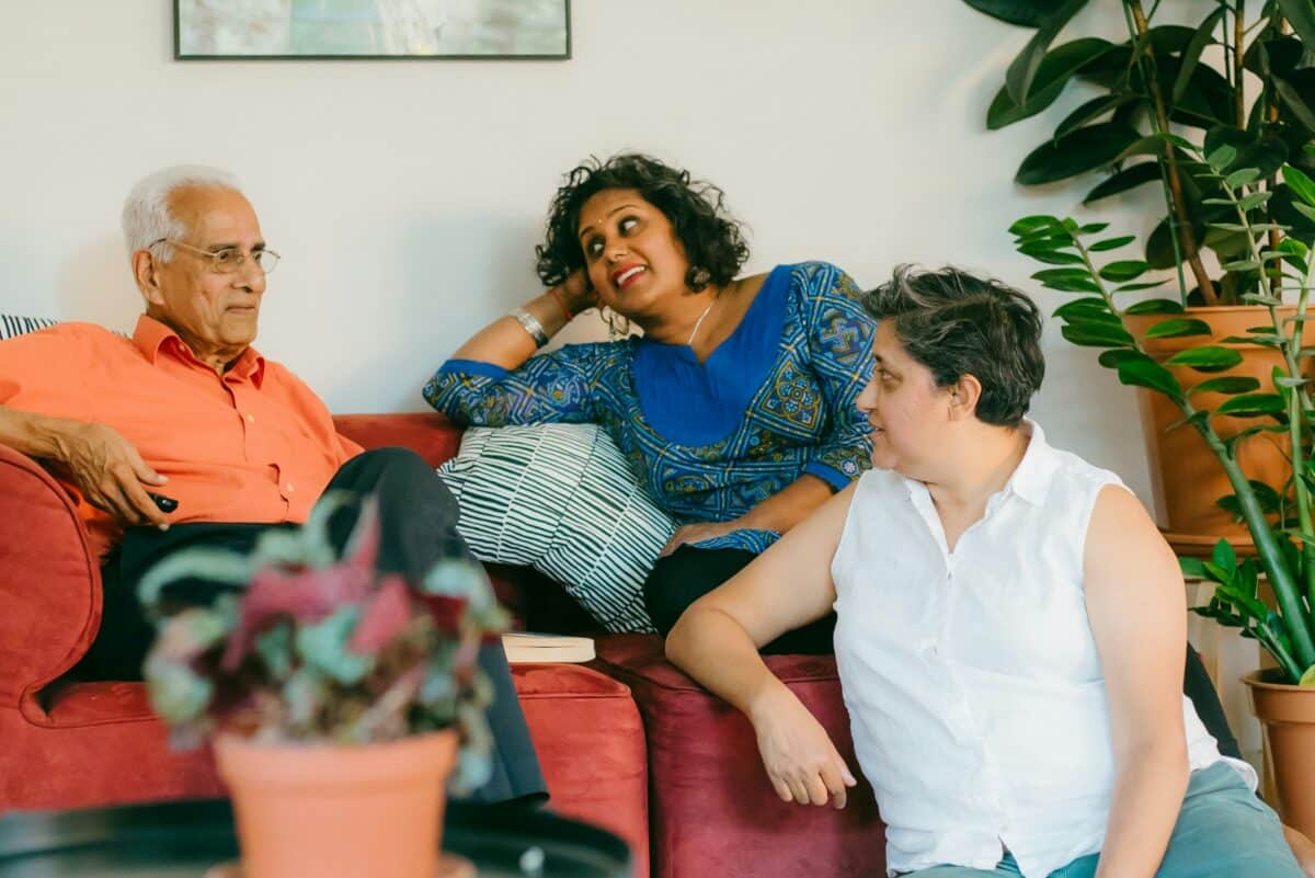An elderly man sitting on a sofa talking and smiling with his adult daughters during a family discussion about moving.