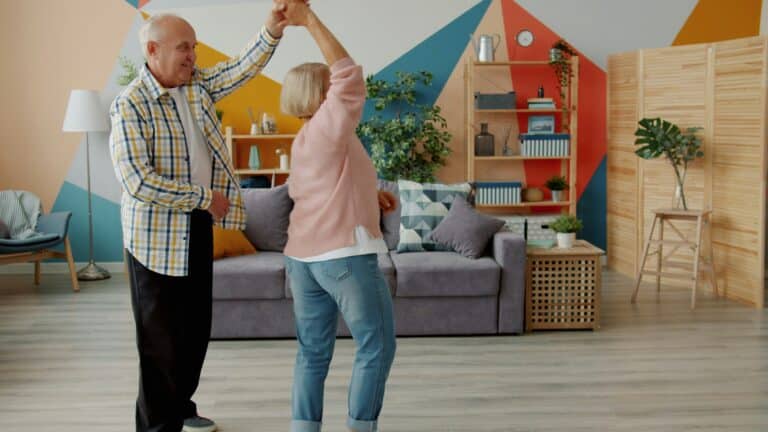 An elderly senior couple smiling and dancing together in a bright, organized living room, illustrating a joyful approach to rightsizing.