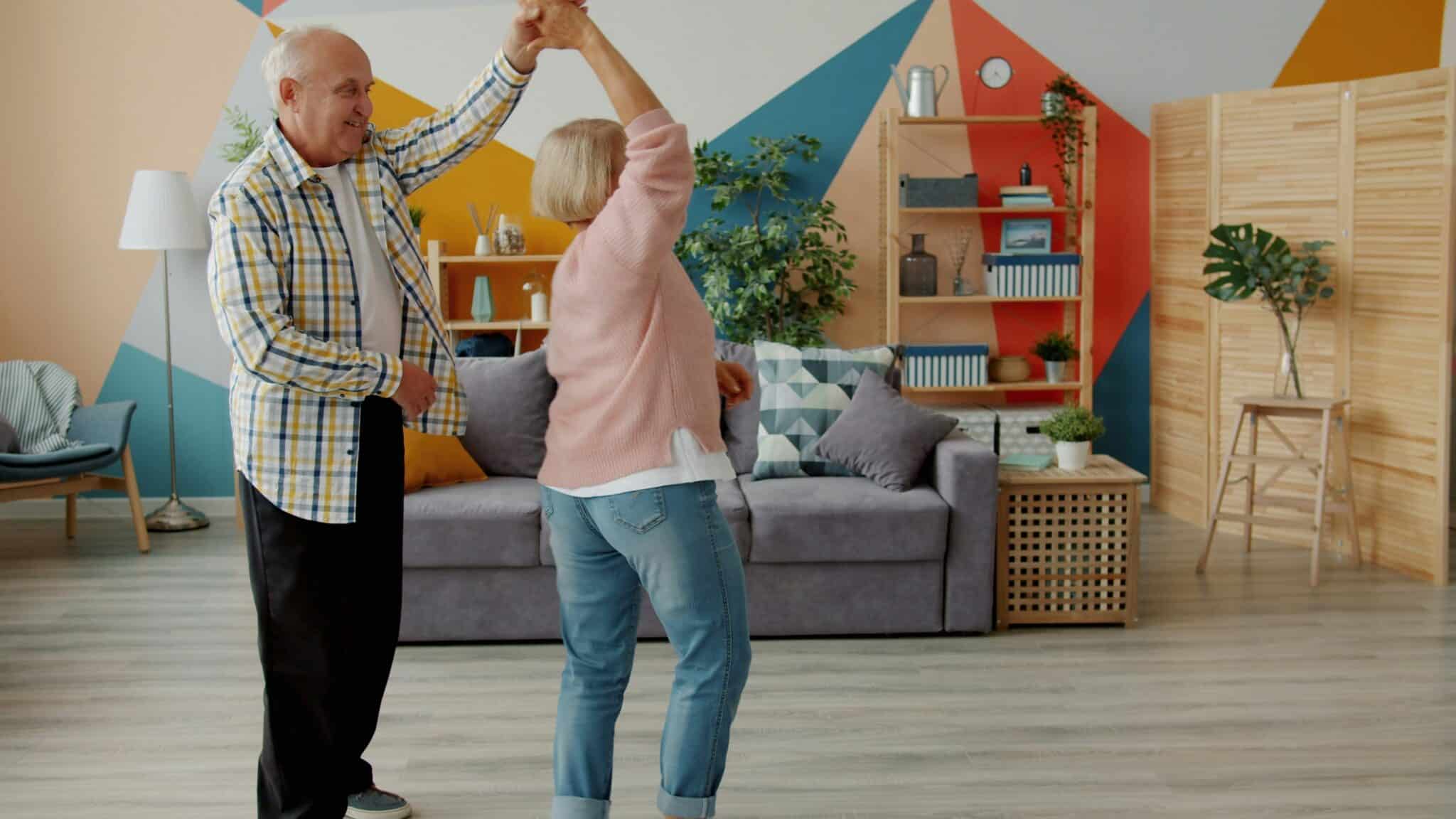 An elderly senior couple smiling and dancing together in a bright, organized living room, illustrating a joyful approach to rightsizing.
