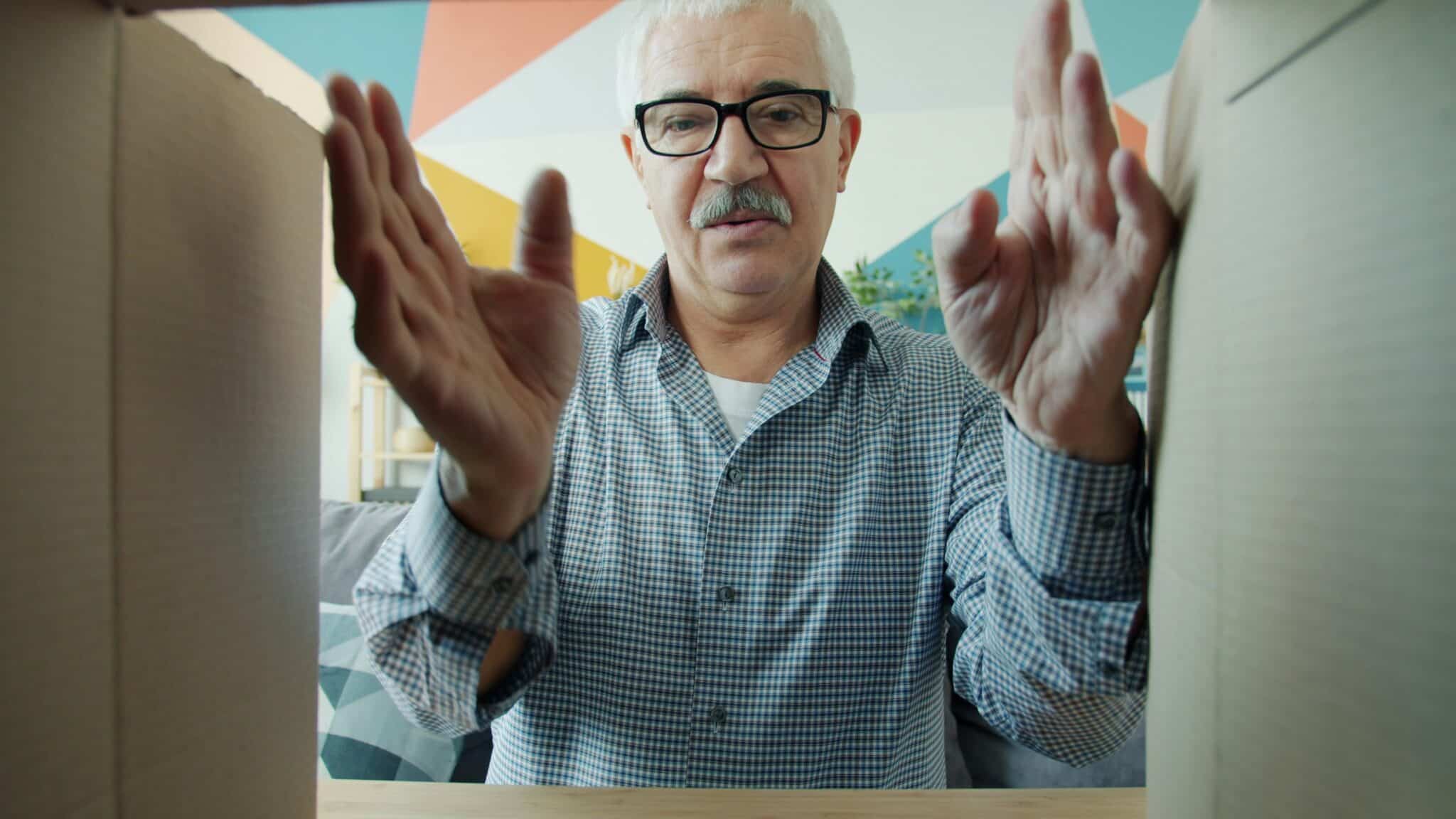 POV shot looking up from inside a cardboard box at a senior man with glasses and a mustache opening and reaching into a moving box.