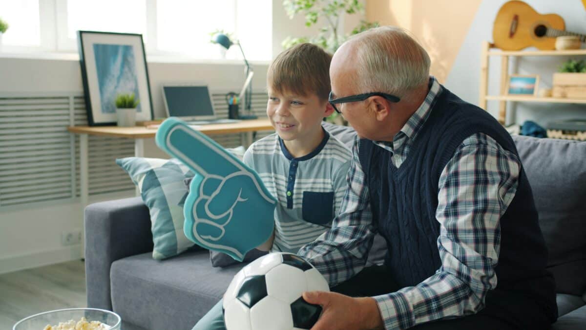 A grandfather sitting on a couch smiling at his young grandson who is holding a soccer ball and a blue foam finger.