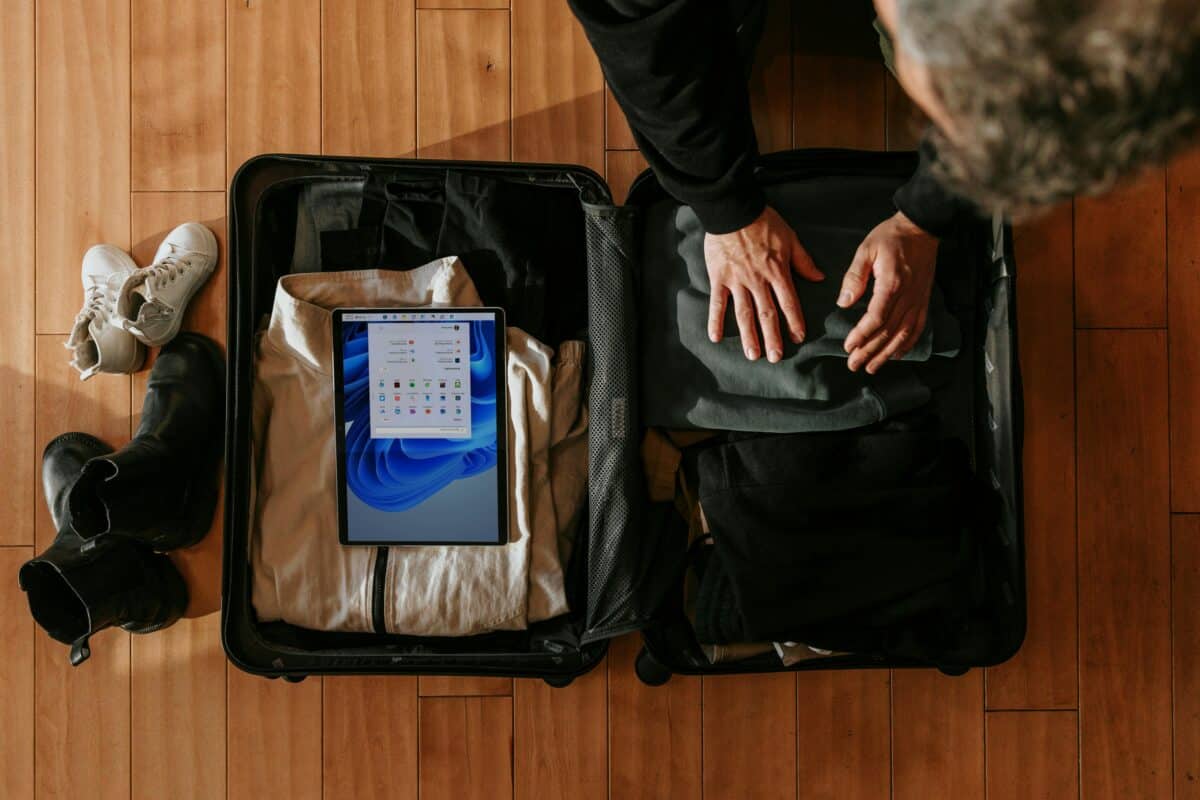 Overhead view of a person packing an open suitcase with folded clothes, shoes, and a tablet device on a wooden floor.