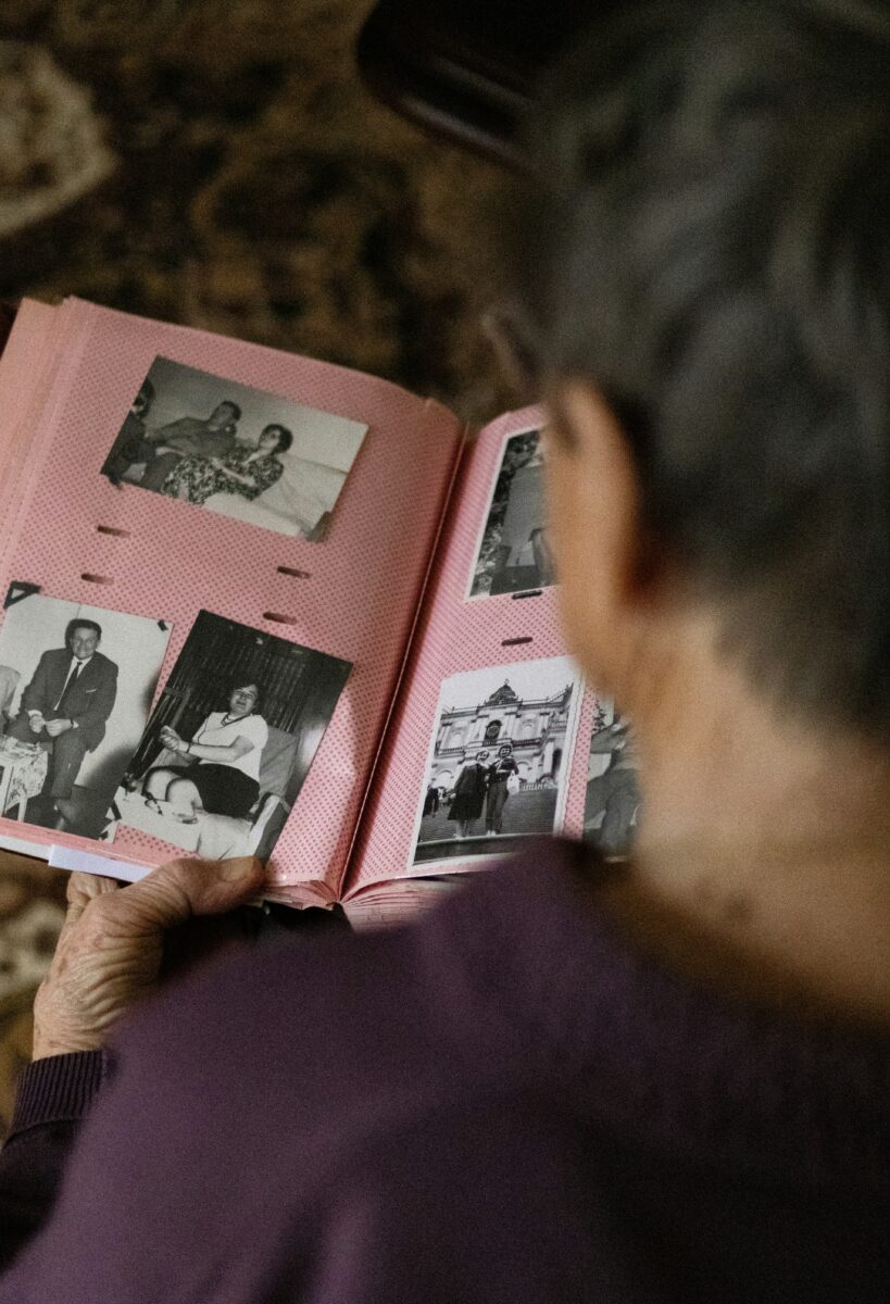 Over-the-shoulder view of an elderly person holding a pink vintage photo album filled with old black-and-white family pictures.